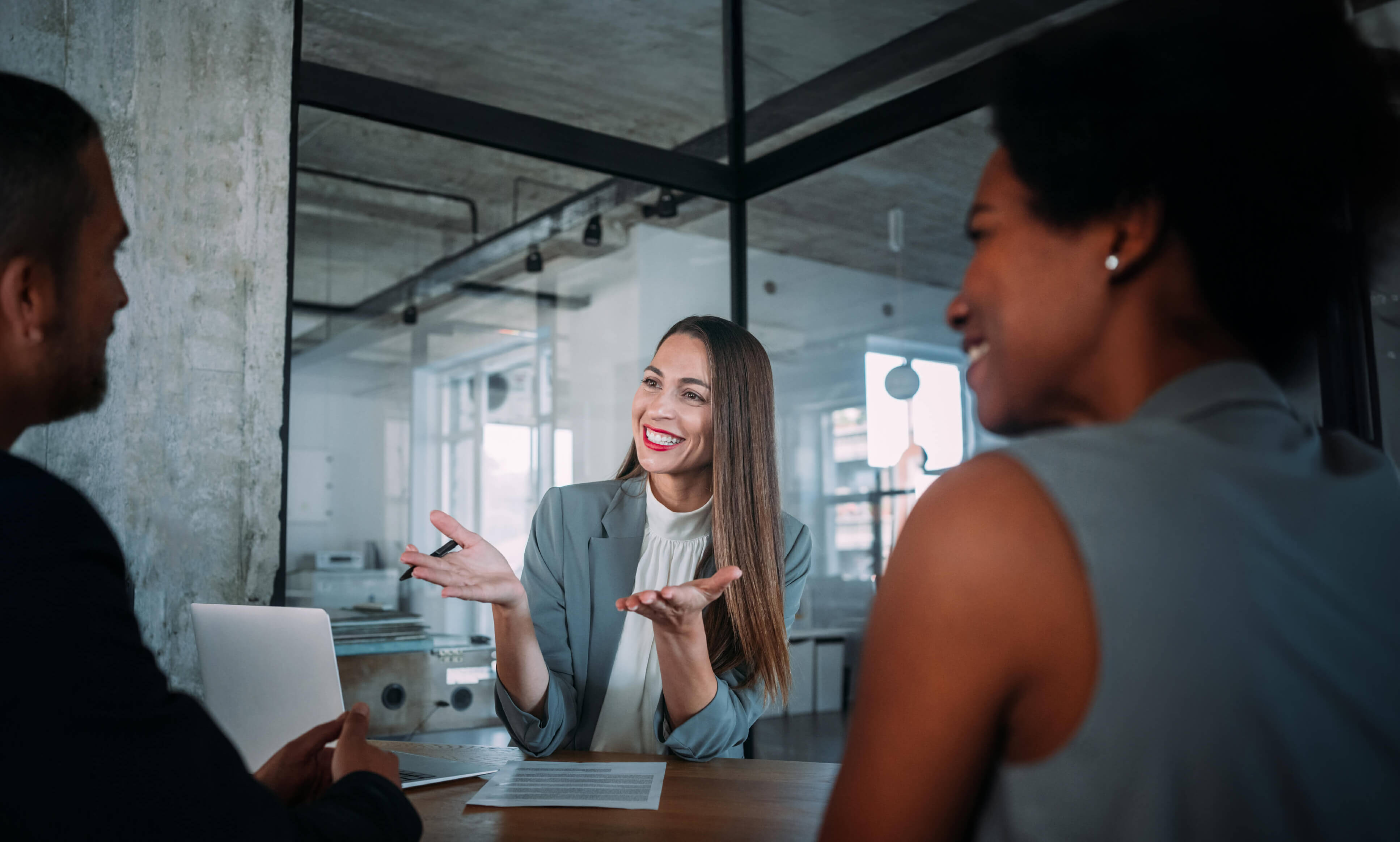 Angeregte Beratungssituation einer jungen, lächelnden Beraterin mit zwei weiteren Personen, die ihr in einem modernen Loft-Büro gegenübersitzen. Marina Adler – Marketing mit Weitblick